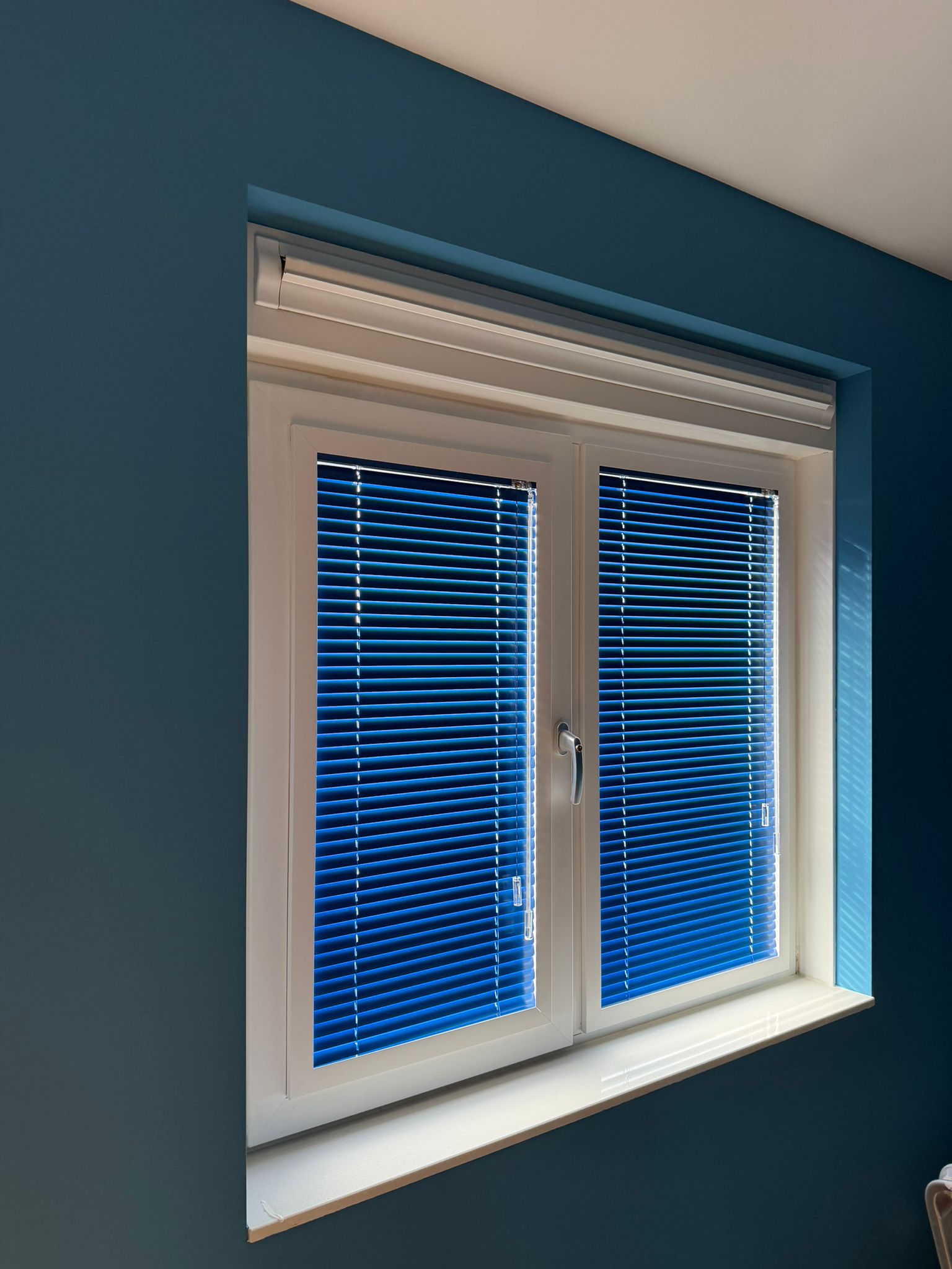 Durf te kiezen voor kleur. Deze kobaltblauwe aluminium jaloezieën geven een krachtig en modern accent aan de donkere muur, terwijl het zonlicht sfeervol wordt gefilterd.