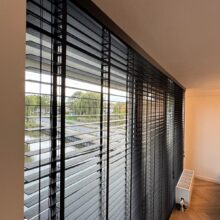 Zwarte houten jaloezieën met ladderband, gemonteerd over de volledige breedte van een raam in een moderne woonkamer.