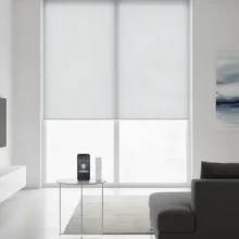 Een modern, lichtgrijs elektrisch rolgordijn dat automatisch naar beneden gaat, met op de voorgrond een smartphone met de bedieningsapp.