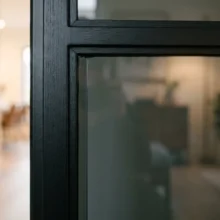 Close-up van een zwarte binnendeur met donker rookglas, waarin het licht subtiel weerspiegelt en de kamer erachter onscherp zichtbaar is.